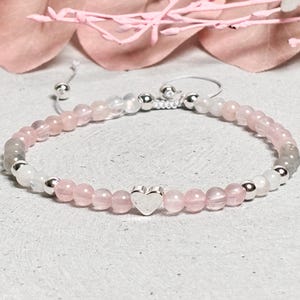 Op de afbeelding: Een armband met roze, witte en grijze edelsteen kralen en een zilveren hartbedel. De kralen zijn geregen op een wit koord en de armband heeft een verstelbare sluiting. Gepresenteerd op een witte, getextureerde ondergrond.
