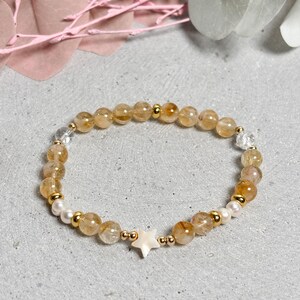 Könnte beinhalten: Ein Armband mit goldgelben und klaren Quarzperlen, unterbrochen von kleinen goldfarbenen Perlen und weißen Perlen. Ein weißer sternförmiger Anhänger befindet sich in der Mitte. Das Armband wird auf einer hellgrauen Oberfläche präsentiert.