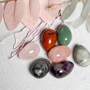 May include: An assortment of polished gemstone eggs in various colors, including rose quartz, green, red, and purple. Decorative pink and white leaves and branches are arranged around the eggs on a light gray surface. The eggs vary in size and texture.