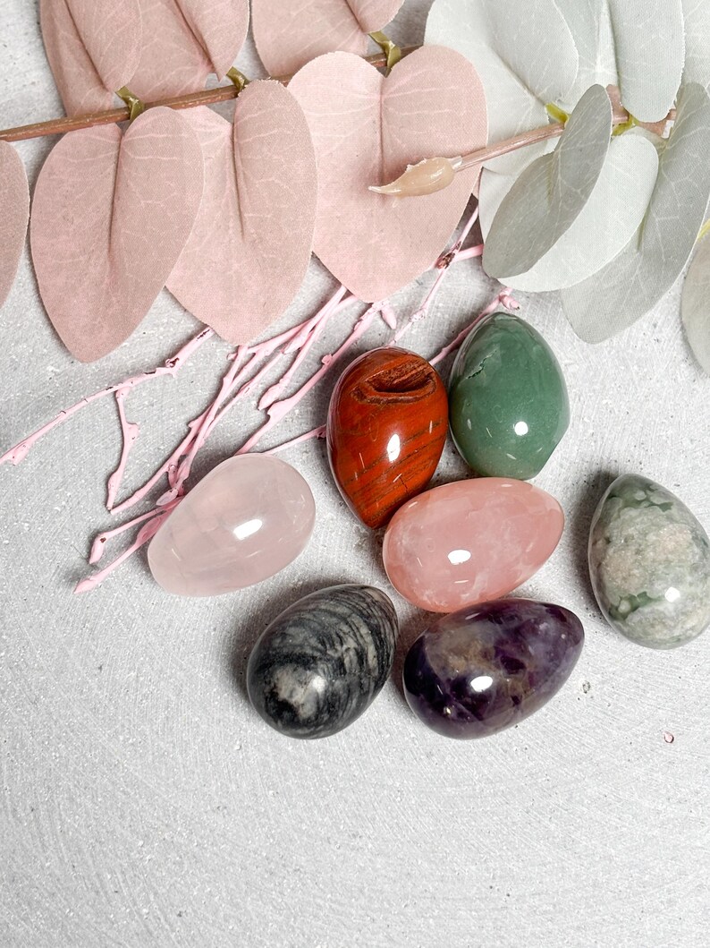 May include: An assortment of polished gemstone eggs in various colors, including rose quartz, green, red, and purple. Decorative pink and white leaves and branches are arranged around the eggs on a light gray surface. The eggs vary in size and texture.