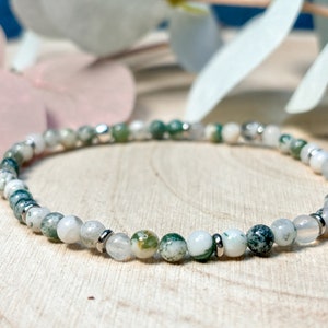 Könnte beinhalten: Ein zartes Perlenarmband mit kleinen, runden, weißen und grünen Moosachatsteinen. Das Armband ist auf einem elastischen Band aufgefädelt und mit silberfarbenen Metallperlen akzentuiert. Die Steine haben ein natürliches, gesprenkeltes Aussehen.