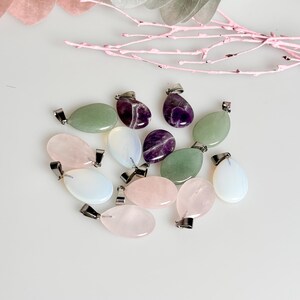 Könnte beinhalten: Eine Sammlung ovaler Edelstein-Anhänger mit silberfarbenen Metallösen. Die Steine sind in Grün-, Rosa-, Weiß- und Lilatönen.