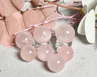 May include: Seven pink rose quartz spheres of varying sizes on a light gray surface. The spheres are smooth and translucent, with a slight pink hue.