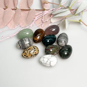 May include: A collection of polished gemstone eggs in various colors and patterns, including green, brown, gray, and white. The eggs are arranged on a white surface with pink and gray decorative branches in the background.
