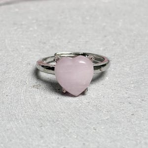 Könnte beinhalten: Verstellbarer Ring in Silberoptik mit einem herzförmigen, hellrosa Edelstein. Der Ring hat ein schlichtes Band, und das Herz ist der Mittelpunkt, vor einem neutralen Hintergrund. Ein zarter Schmuck.