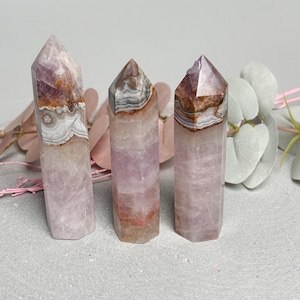 Könnte beinhalten: Drei Amethyst-Kristalltürme mit unterschiedlichen Mustern und Farben. Die Kristalle sind hauptsächlich in Lila gehalten, mit Bändern aus Weiß, Braun und Orange an der Spitze. Die Kristalle werden auf einer grauen Oberfläche präsentiert.