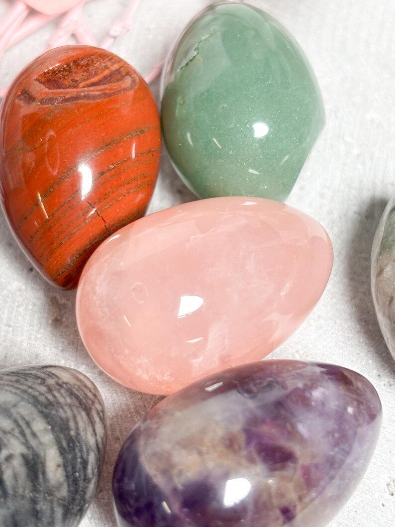 May include: Assortment of polished gemstone eggs in various colors. Includes a red-orange jasper egg, a light green aventurine egg, a pink rose quartz egg, and a purple amethyst egg. The eggs are smooth and have a glossy finish.