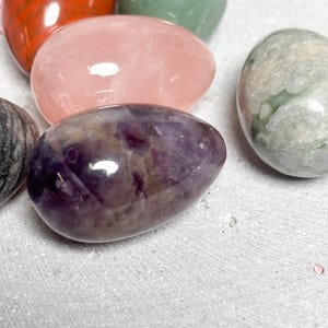 May include: A collection of polished gemstone eggs in various colors. The eggs include red, green, pink, purple, and gray. The gemstones have a smooth, glossy finish and are arranged on a light gray surface.