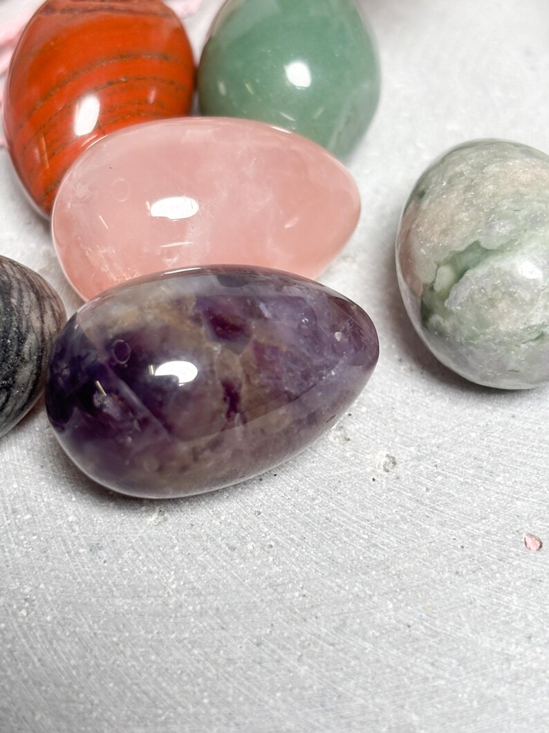May include: A collection of polished gemstone eggs in various colors. The eggs include red, green, pink, purple, and gray. The gemstones have a smooth, glossy finish and are arranged on a light gray surface.