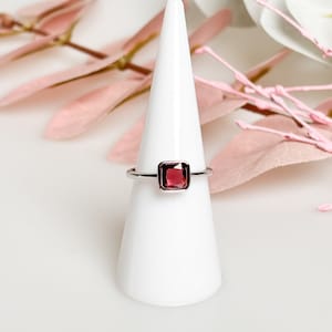 May include: A silver ring with a square, dark red gemstone. The ring is displayed on a white cone-shaped ring holder.