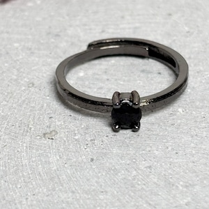 Peut inclure: Une bague noire réglable avec une pierre noire carrée. La bague est posée sur une surface grise avec un objet rose flou en arrière-plan. Le corps de la bague est de couleur canon de fusil foncé.