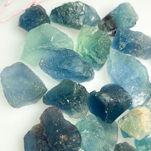 May include: A selection of raw fluorite crystals in various shades of blue and green. The crystals have a rough, textured appearance and are of varying sizes. Some pink twigs are visible in the background.