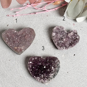 Könnte beinhalten: Drei herzförmige Amethyst-Kristalle auf einer grauen Oberfläche. Die Kristalle sind unterschiedlich groß und haben verschiedene Lilatöne.
