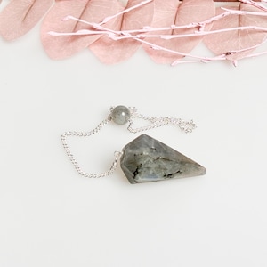 Op de afbeelding: Een zilveren ketting met een grijs kristallen slinger. De slinger heeft een puntige, driehoekige vorm.