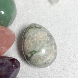 May include: An assortment of polished gemstone eggs in various colors. The central egg is a mottled green and gray, with other eggs in shades of green, pink, and purple. The gemstones are smooth and reflective, suggesting a natural, organic theme.