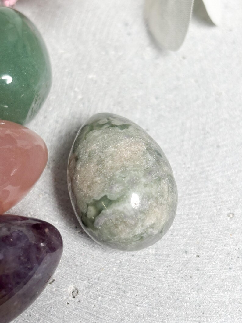 May include: An assortment of polished gemstone eggs in various colors. The central egg is a mottled green and gray, with other eggs in shades of green, pink, and purple. The gemstones are smooth and reflective, suggesting a natural, organic theme.