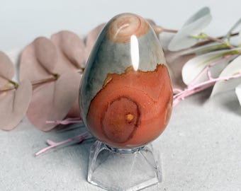 May include: A polished, egg-shaped stone featuring a smooth surface with distinct color bands of muted gray, terracotta orange, and cream. The stone is displayed upright on a clear, faceted hexagonal plastic stand for stability.