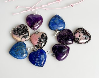 May include: A collection of heart-shaped gemstone pendants in various colors, including deep purple, blue, and pink. Each pendant has a silver bail for attaching to a chain. The gemstones have unique patterns and textures.