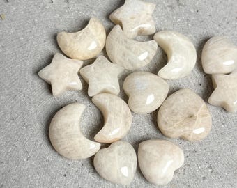 May include: A collection of small, polished moonstone carvings in various shapes, including crescent moons, five-pointed stars, and hearts. Each piece features a smooth, glossy surface with a creamy, off-white color and subtle internal light reflections.