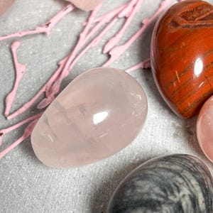 May include: A collection of polished gemstone eggs in various colors. A light pink rose quartz egg is in the foreground, with a red-brown jasper heart-shaped stone and a gray and white patterned egg visible.
