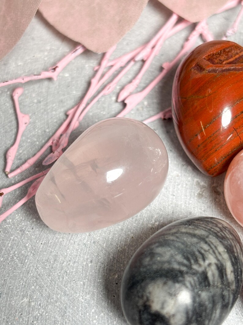 May include: A collection of polished gemstone eggs in various colors. A light pink rose quartz egg is in the foreground, with a red-brown jasper heart-shaped stone and a gray and white patterned egg visible.