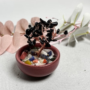Puede incluir: Una escultura de árbol decorativa con ramas de alambre de cobre trenzado adornadas con pequeñas piedras negras pulidas. El árbol está plantado en un pequeño cuenco de cerámica de color rojo oscuro lleno de guijarros de piedras preciosas.