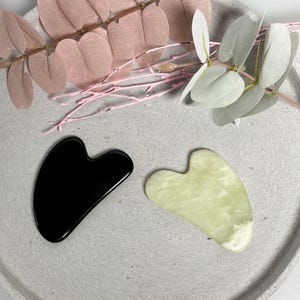 Könnte beinhalten: Zwei Gua-Sha-Werkzeuge in Herzform, eines schwarz und eines hellgrün, liegen auf einem grauen, kreisförmigen Tablett. Die Werkzeuge haben glatte, polierte Oberflächen. Rosa und weiße dekorative Blätter und Zweige sind im Hintergrund.