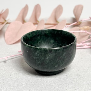 Könnte beinhalten: Eine kleine, dunkelgrüne Steinschale mit abgerundeter Form. Die Schale hat eine glatte, polierte Oberfläche und steht auf einem flachen Sockel. Der Hintergrund zeigt rosa florale Akzente, die einen Kontrast zur Farbe der Schale bilden.