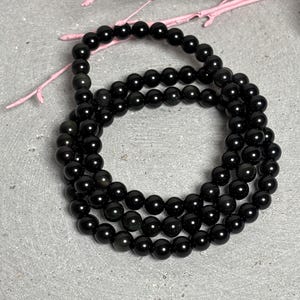 Può includere: Una collana di perline nere, composta da numerose piccole perline rotonde. La collana è arrotolata e poggia su una superficie grigia, con un accento floreale rosa sullo sfondo. Le perline hanno una finitura lucida, riflettendo la luce.