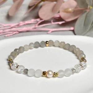 May include: A bracelet with grey, white, and clear beads, with small gold-coloured spacers. The design includes a central pearl. The bracelet is presented on a white surface.