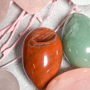 May include: Close-up of polished gemstone eggs in shades of red, green, and pink. The red egg has brown striations. Decorative pink branches and a light gray surface complete the composition. These eggs are ideal for home decor or as a unique gift.