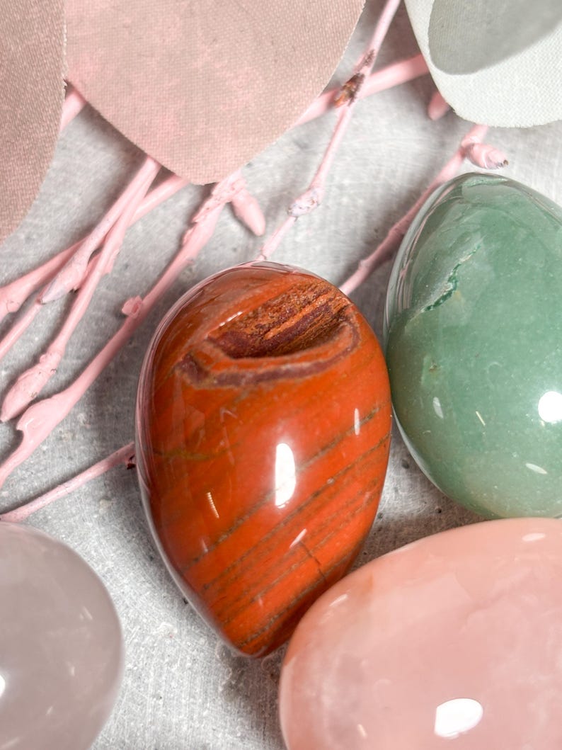 May include: Close-up of polished gemstone eggs in shades of red, green, and pink. The red egg has brown striations. Decorative pink branches and a light gray surface complete the composition. These eggs are ideal for home decor or as a unique gift.