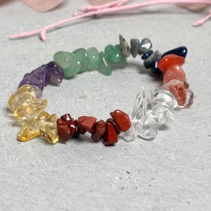 Könnte beinhalten: Ein farbenfrohes Armband aus polierten Edelsteinen. Das Armband weist eine Vielzahl von Farben auf, darunter Lila, Gelb, Grün, Rot und Klar. Die Steine sind unregelmäßig geformt und miteinander aufgereiht, wodurch ein einzigartiges Accessoire entsteht.
