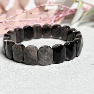Könnte beinhalten: Ein dunkelgraues und schwarzes Armband aus rechteckigen, polierten Steinen. Das Armband wird auf einer weißen Oberfläche mit rosa floralen Akzenten im Hintergrund gezeigt. Die Steine haben ein marmoriertes Aussehen.