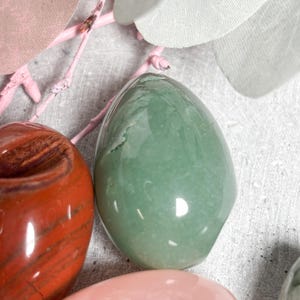 May include: A collection of polished gemstones in various colors, including green, red, and pink. The smooth, egg-shaped stones are displayed with pink branches and light-colored fabric leaves, creating a natural and serene aesthetic.
