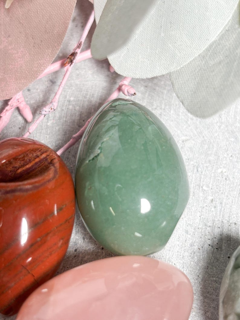 May include: A collection of polished gemstones in various colors, including green, red, and pink. The smooth, egg-shaped stones are displayed with pink branches and light-colored fabric leaves, creating a natural and serene aesthetic.