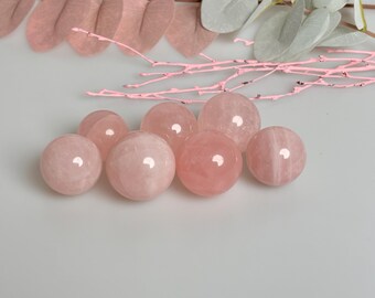 May include: A group of seven pink rose quartz spheres, some with visible striations, arranged on a white surface.
