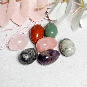 Peut inclure: Un ensemble de sept œufs polis en pierres précieuses de différentes couleurs, dont rose, rouge, vert, gris et violet. Les œufs sont disposés sur une surface gris clair avec des éléments floraux roses et blancs.