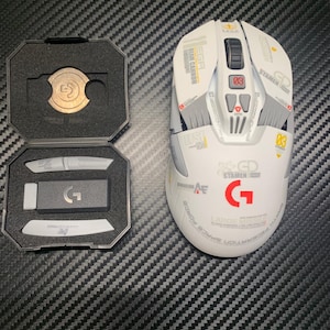 May include: A white and gray gaming mouse with red and yellow accents, and the letter "G" in red. The mouse is next to an open black case containing a gold-colored coin and other accessories. The text "EGA", "STAMEN", and "LARGE MISSILE" are visible.