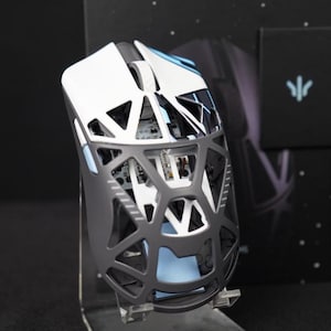 Könnte beinhalten: Eine Gaming-Maus in Weiß und Schwarz. Die Maus hat ein einzigartiges Open-Frame-Design mit einem geometrischen Muster. Sie wird auf einem transparenten Ständer präsentiert, mit einer schwarzen Box und einem Logo im Hintergrund.