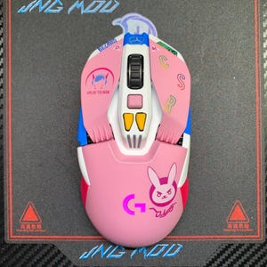 Può includere: Un mouse da gioco rosa e bianco con accenti blu e gialli. Il mouse presenta il testo "I PLAY TO WIN" e una grafica a forma di coniglietto. Il mouse è su una superficie nera con la scritta "JNG MOD".