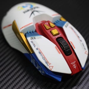 Puede incluir: Un ratón para juegos blanco y rojo con detalles dorados, azules y amarillos. El ratón tiene una rueda de desplazamiento, botones y texto que dice "REMOVABLE TANK" y "102". El diseño recuerda a un mecha.
