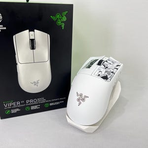 May include: A white Razer Viper V3 Pro gaming mouse with a white charging dock. The mouse has a black scroll wheel and a black and white graphic design on the top. The box is black with the Razer logo and product information.