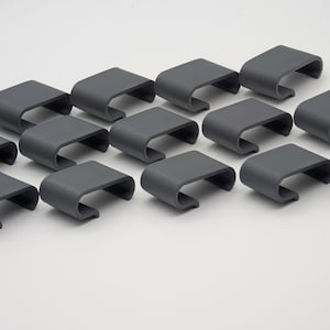 Könnte beinhalten: Eine Ansammlung von sechzehn grauen, gebogenen, rechteckigen Clips, die auf einer weißen Oberfläche angeordnet sind. Jeder Clip hat eine glatte, matte Oberfläche und ein schlichtes, modernes Design. Die Clips sind gleichmäßig verteilt.