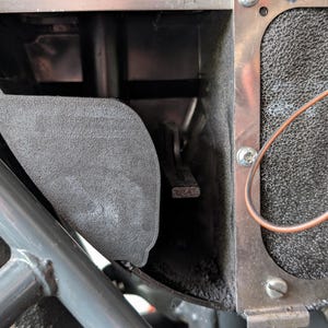 Könnte beinhalten: Nahaufnahme einer Motorradkomponente mit grauem Metallrahmen und strukturiertem, dunkelgrauem Paneel. Ein kupferfarbener Draht ist sichtbar, zusammen mit einem Teil eines Reifens. Das Design deutet auf einen Fokus auf mechanische Teile und Technik hin.