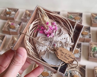 May include: A small, white, round soap bar wrapped in burlap and lace, with dried pink flowers and a wooden tag that reads "Merve & Davut 05.05.2020" in a clear box with a straw nest.