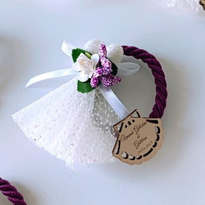 May include: A white tulle favor bag with a purple flower and a white ribbon. The bag is tied with a purple rope and has a wooden tag with the text "Ummi Gülşüm & Gökhan 19.04.2023" in brown lettering.