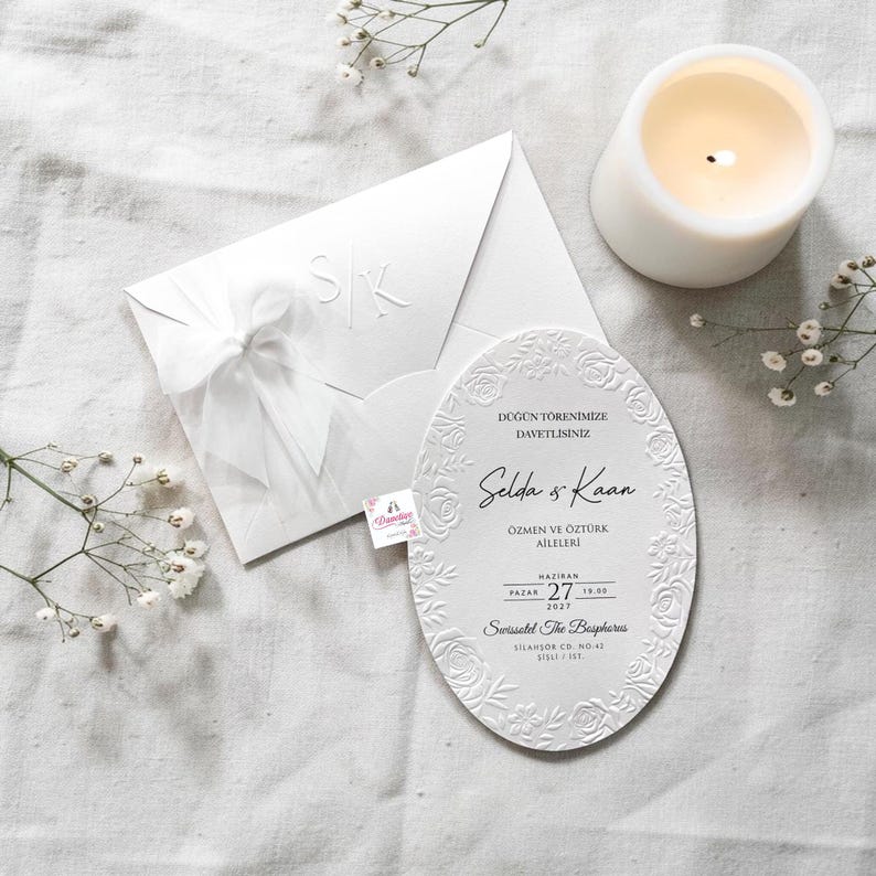 May include: A white wedding invitation suite with an embossed floral design. The suite includes an envelope with initials and a ribbon, and an oval invitation card with text. A lit candle and sprigs of white flowers are also present.