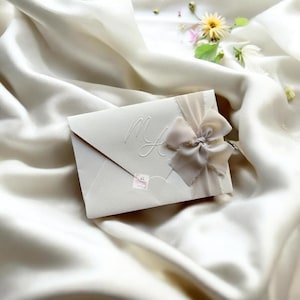 Pode incluir: Envelope cor marfim com iniciais em relevo e um delicado laço de fita. O envelope está sobre um fundo de tecido de cetim branco, com algumas flores pequenas ao fundo. A estética geral é elegante e refinada.