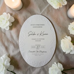 May include: An oval-shaped wedding invitation with embossed floral details and text. The invitation features the names "Selda & Kaan", event details, and the venue "Swissotel The Bosphorus". White flowers and candles surround the invitation.